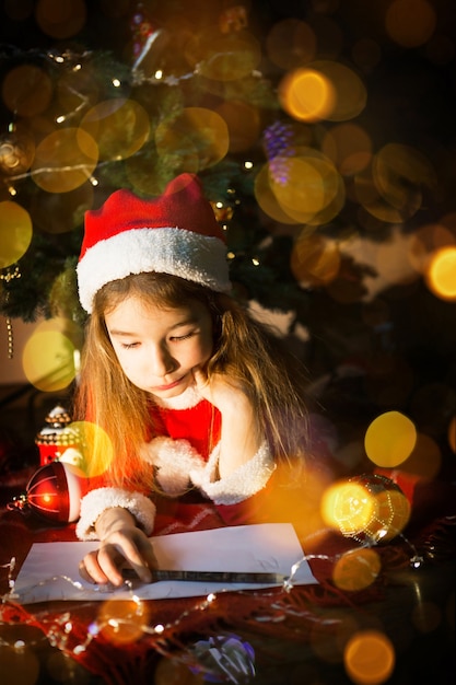 Little girl in a Santa hat and red dress under Christmas tree is dreaming, waiting for the holiday, lying on a plaid blanket. A letter on piece of paper, gifts. New year, Christmas. defocus lights