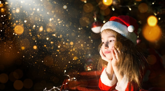 Little girl in a Santa hat and red dress under Christmas tree is dreaming, waiting for the holiday, lying on a plaid blanket. A letter on piece of paper, gifts. New year, Christmas. defocus lights