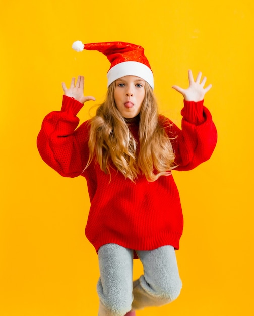 Little girl in a santa hat and a red sweater jumps and shows five on a yellow background.