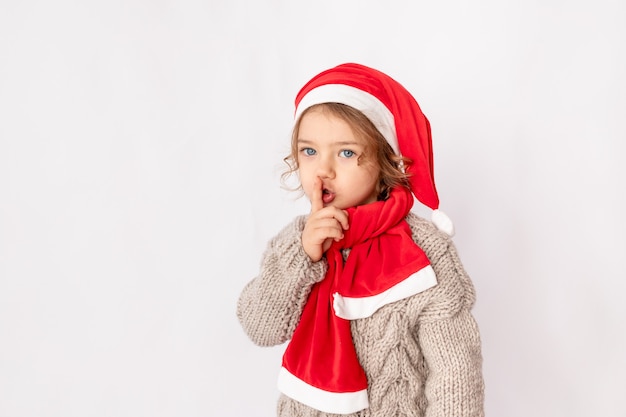 A little girl in a Santa hat shows her finger quietly on a white background, space for text