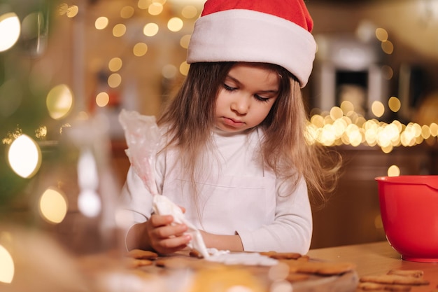 Little girl in santas hat decorates gingerbread using white glaze christmas and new year traditions