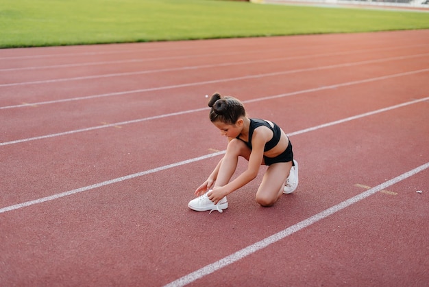 Маленькая девочка села завязывать шнурки перед беговой тренировкой на стадионе во время заката Детский спорт и здоровый образ жизни