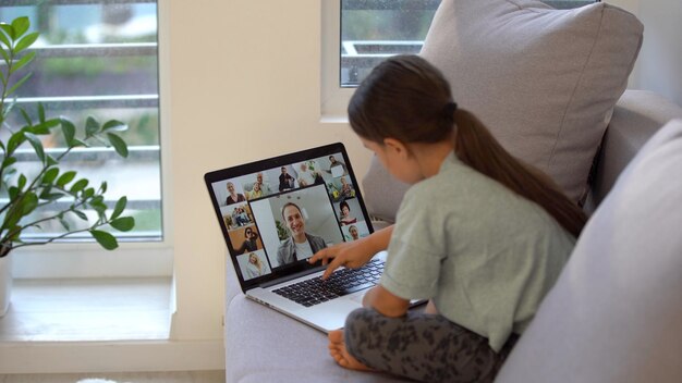 Little girl of school age is studying at home. a girl watches a\
video lesson on a laptop monitor and writes tasks in a\
notebook.