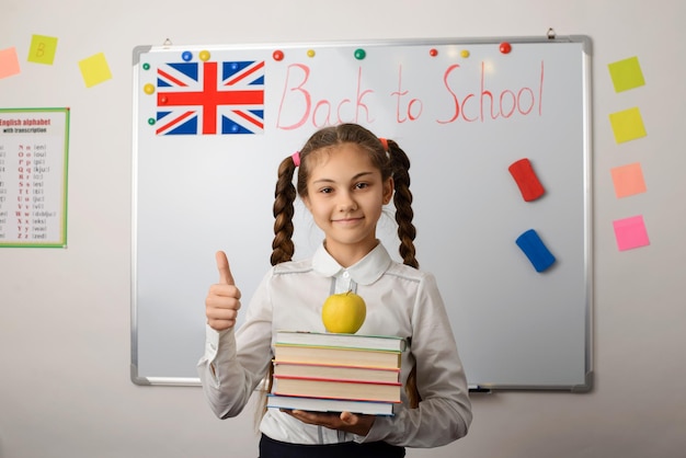 Bambina in uniforme scolastica con una pila di libri vicino alla lavagna a scuola che mostra i pollici in su