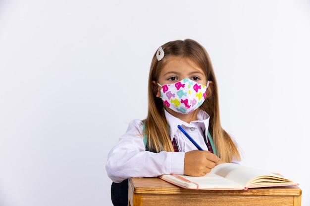 Bambina a scuola che indossa una maschera protettiva per proteggersi, seduta accanto a un tavolo con