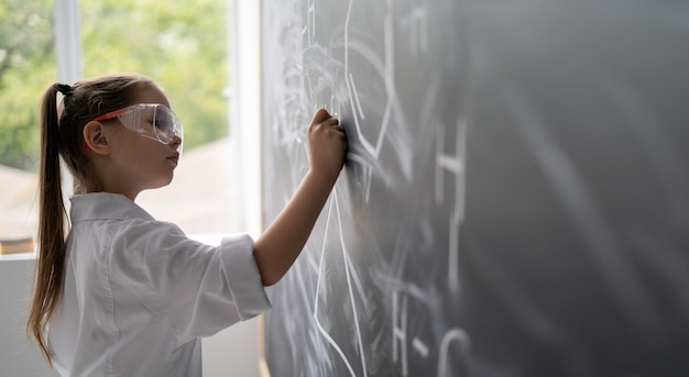 黒板の近くの小さな女の子の女子高生は、化学の問題の白衣とゴーグルを解決します