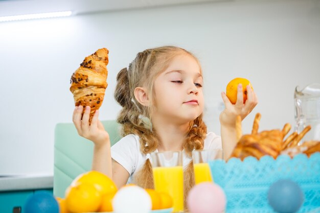 A little girl of seven, wearing a white T-shirt, sits at the kitchen table. Holds mandarine tangerines and a freshly baked croissant with chocolate