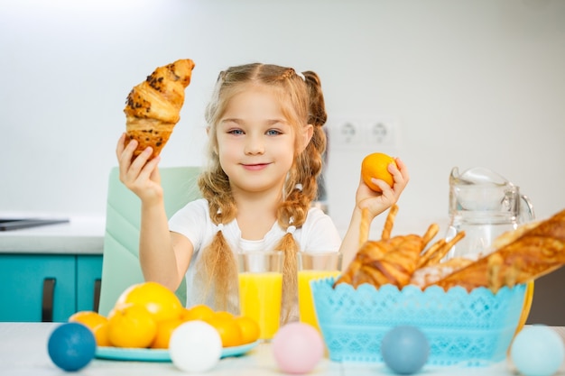 Una bambina di sette anni, con indosso una maglietta bianca, è seduta al tavolo della cucina. contiene mandarini mandarini e un croissant appena sfornato al cioccolato