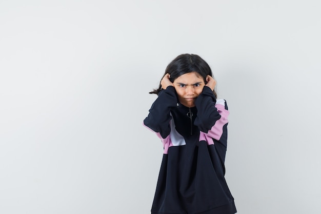 Little girl in shirt holding hands on ears and looking scared , front view.