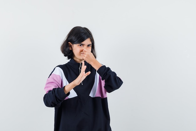 Little girl in shirt pinching nose due to bad smell and looking disgusted , front view.