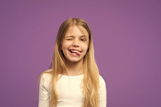 Little girl show tongue Child with funny face on violet background Cute kid with beauty look and long blond hair Beauty look and hair care Happy childhood and childcare copy space