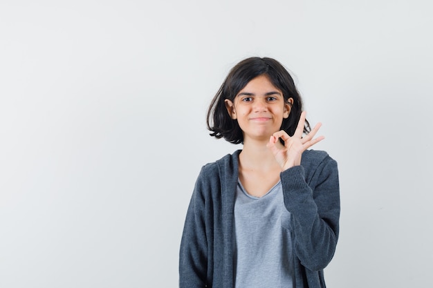 Little girl showing ok gesture in t-shirt, jacket and looking merry , front view.