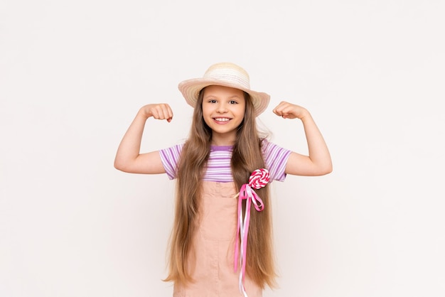 A little girl shows her biceps and holds a caramel on a stick in her pocket on a white isolated background