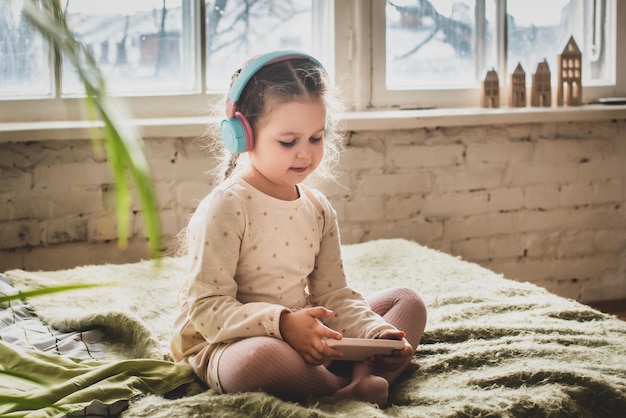 A little girl sits on the bed in headphones baby recklessly plays games and listens to music