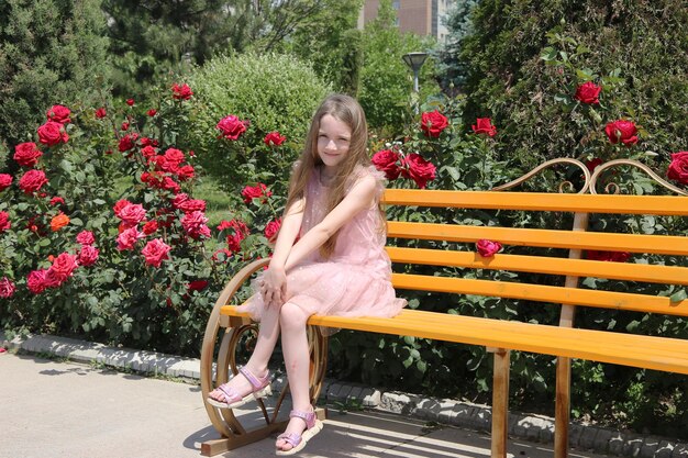 A little girl sits on a bench among red roses