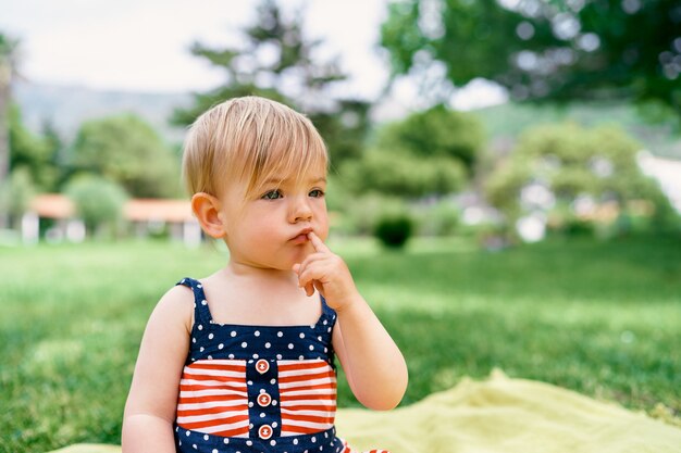 Little girl sits on a blanket on the lawn and holds her finger near her lips