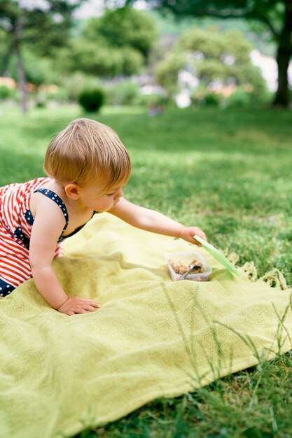 Little girl sits on a blanket on the lawn and reaches for a plastic container with food