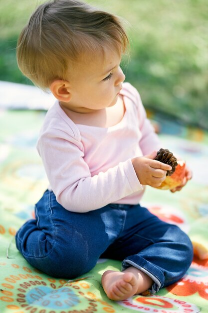 Photo little girl sits on a colored blanket and holds a pine cone and an apple in her hands