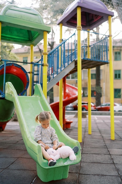 Little girl sits on a colorful slide at the playground and plays with a plush bunny