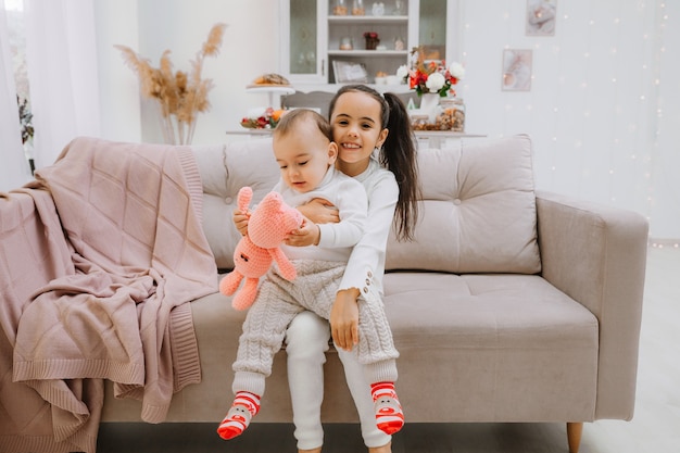 A little girl sits on the couch and holds her little brother on her lap.