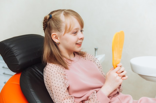 A little girl sits in a dental chair and examines her cured teeth while looking in the mirror