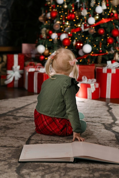 Little girl sits on the floor with large book and looks at\
beautiful christmas tree back view vertical frame