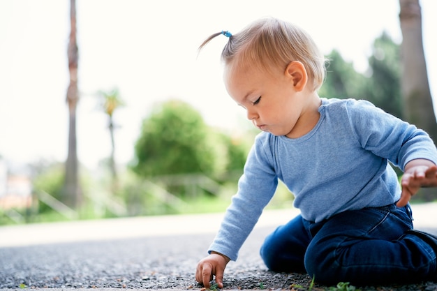 Little girl sits on her lap and picks the asphalt in a green park side view