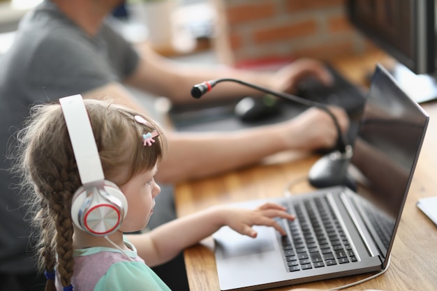 Little girl sits at laptop in headphones with microphone next to man