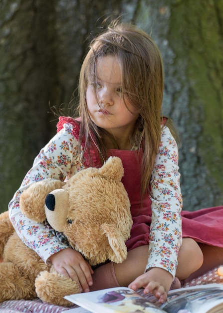 A little girl sits near a large tree in the park holding a teddy bear in her hands and looking away