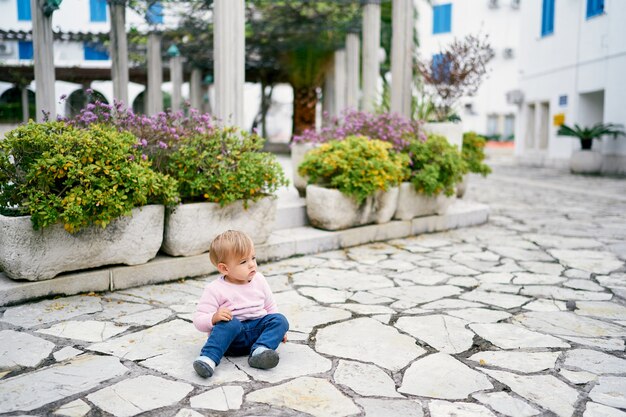 La bambina si siede sulle lastre per pavimentazione vicino ai vasi da fiori con i cespugli verdi sui precedenti degli edifici