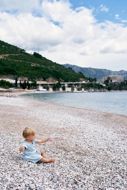 Little girl sits on a pebble beach by the sea with a bottle of water in her hand