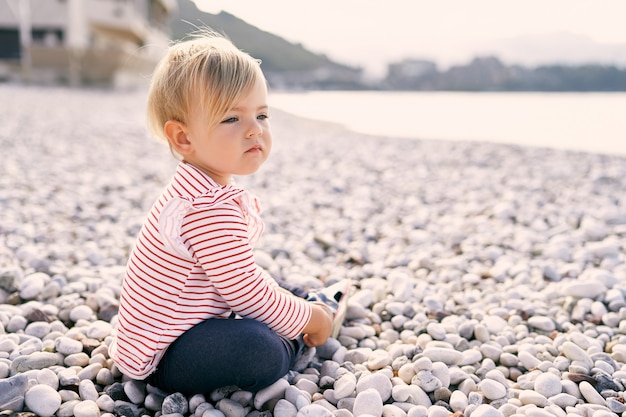 어린 소녀는 자갈 해변에 앉아 바다 쪽 전망을 본다