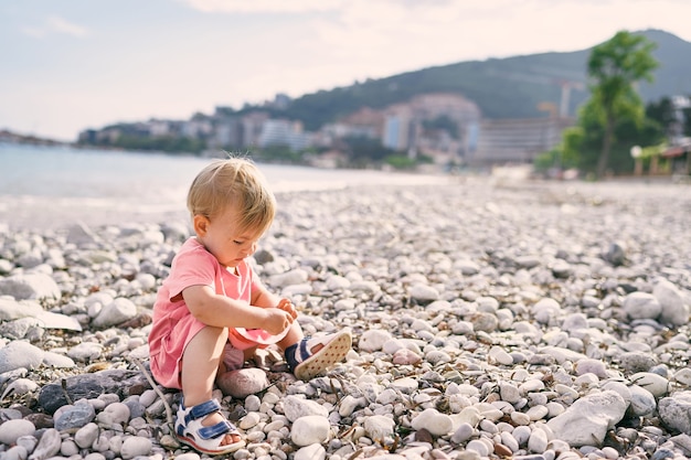 La bambina si siede su una spiaggia di ciottoli e gioca con i ciottoli