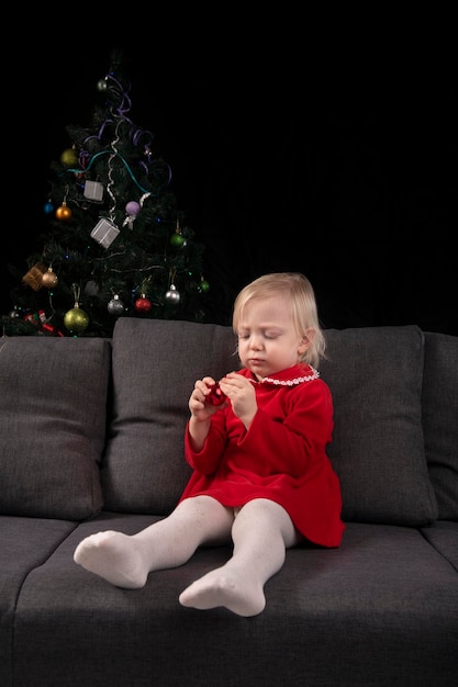 Little girl sits on a sofa against the background of a Christmas tree and looks at a Christmas tree toy New Year eve Vertical frame