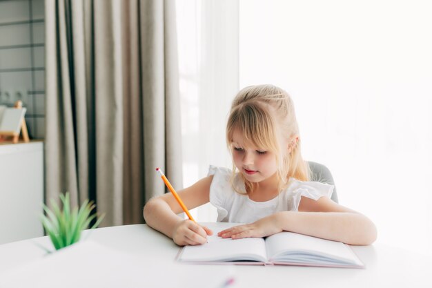 A little girl sits at the table and writes in a white notebook