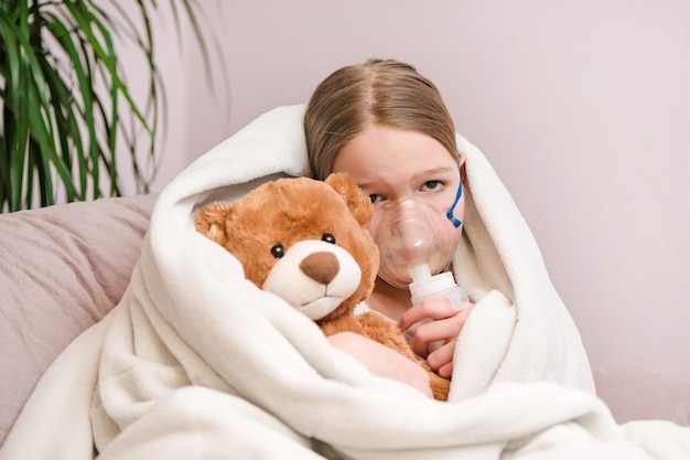 Little girl Sits with toy on sofa at home in a mask for inhalations, making inhalation with nebulizer at home inhaler.