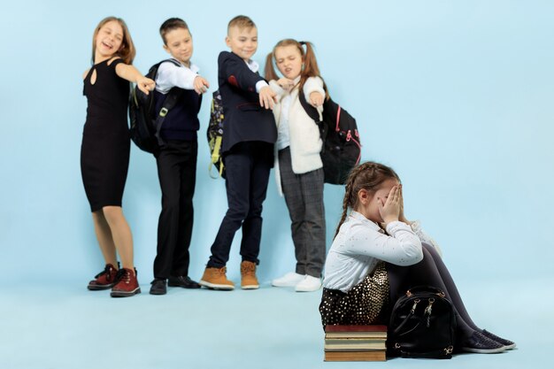 Little girl sitting alone on floor and suffering an act of bullying while children mocking. Sad young schoolgirl sitting on studio against blue background.