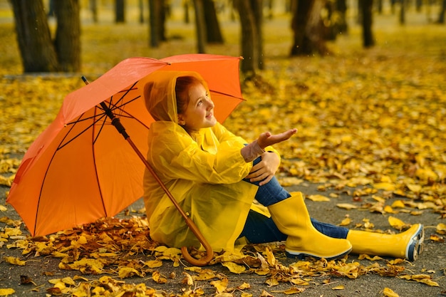 Little girl sitting on the asphalt under an orange umbrella in the rain. Autumn outdoor fun for children. Kid enjoying autumn rain.