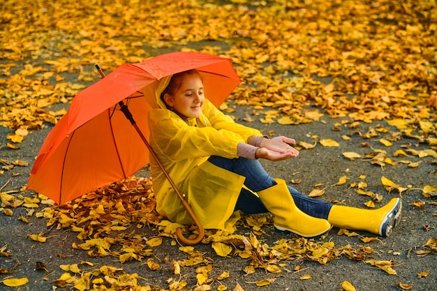 雨の中でオレンジ色の傘の下でアスファルトに座っている少女。子供のための秋のアウトドアの楽しみ。秋の雨を楽しんでいる子供。