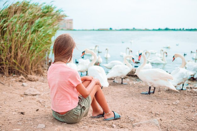 Little girl sitting on the beach with swans and feeding them