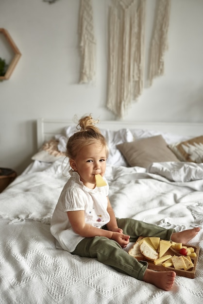 Photo little girl sitting on the bed in the bedroom and eating a piece of cheese.