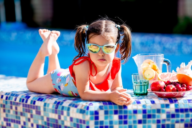 Маленькая девочка сидит у бассейна, пьет лимонад