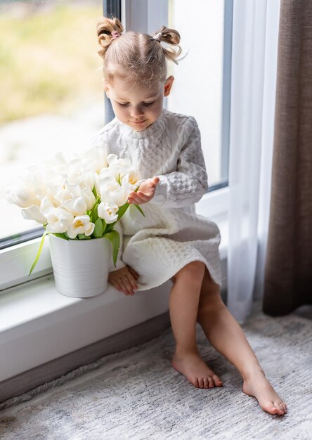 Photo little girl sitting by window with tulip flowers bouquet happy child indoors mothers day valentines day or birthday concept high quality photo