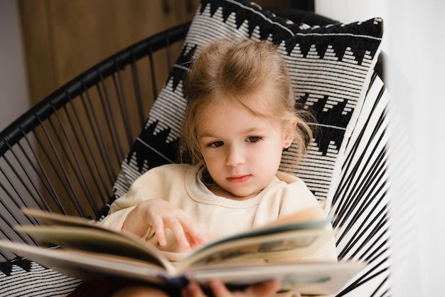 Little girl sitting in a chair by the window reading a book