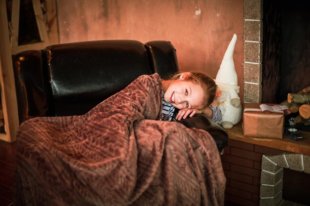 Little girl sitting on cozy chair near christmas tree in morning