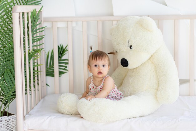 Little girl sitting in a crib at home with a big teddy bear