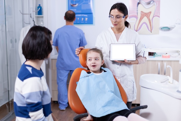 Bambina seduta in poltrona odontoiatrica e dentista pediatrico utilizzando tablet pc con chiave di crominanza. stomatolog che spiega la prevenzione dei denti alla madre e al bambino che tengono tablet pc con copia spazio disponibile.