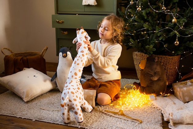 Little girl sitting on floor in Christmas home near festive garland and playing with her cute toy giraffe