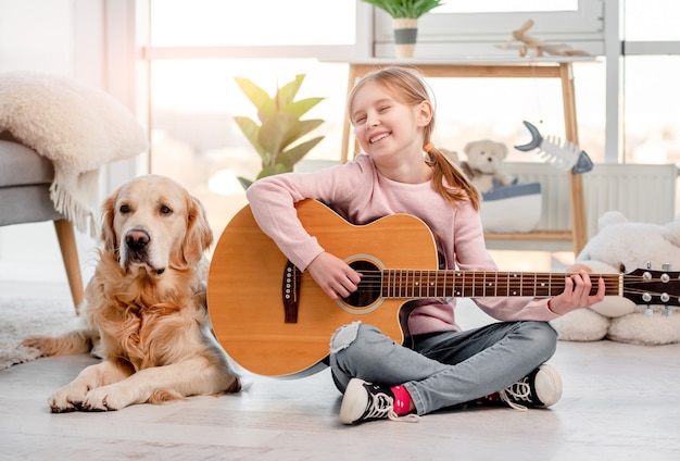 Little girl sitting on the floor and playing guitar with golden retriever dog lying next to her