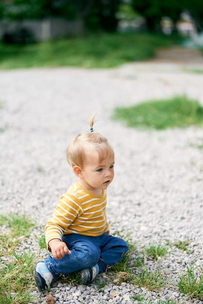 Little girl sitting on a gravel path among the bushes of grass